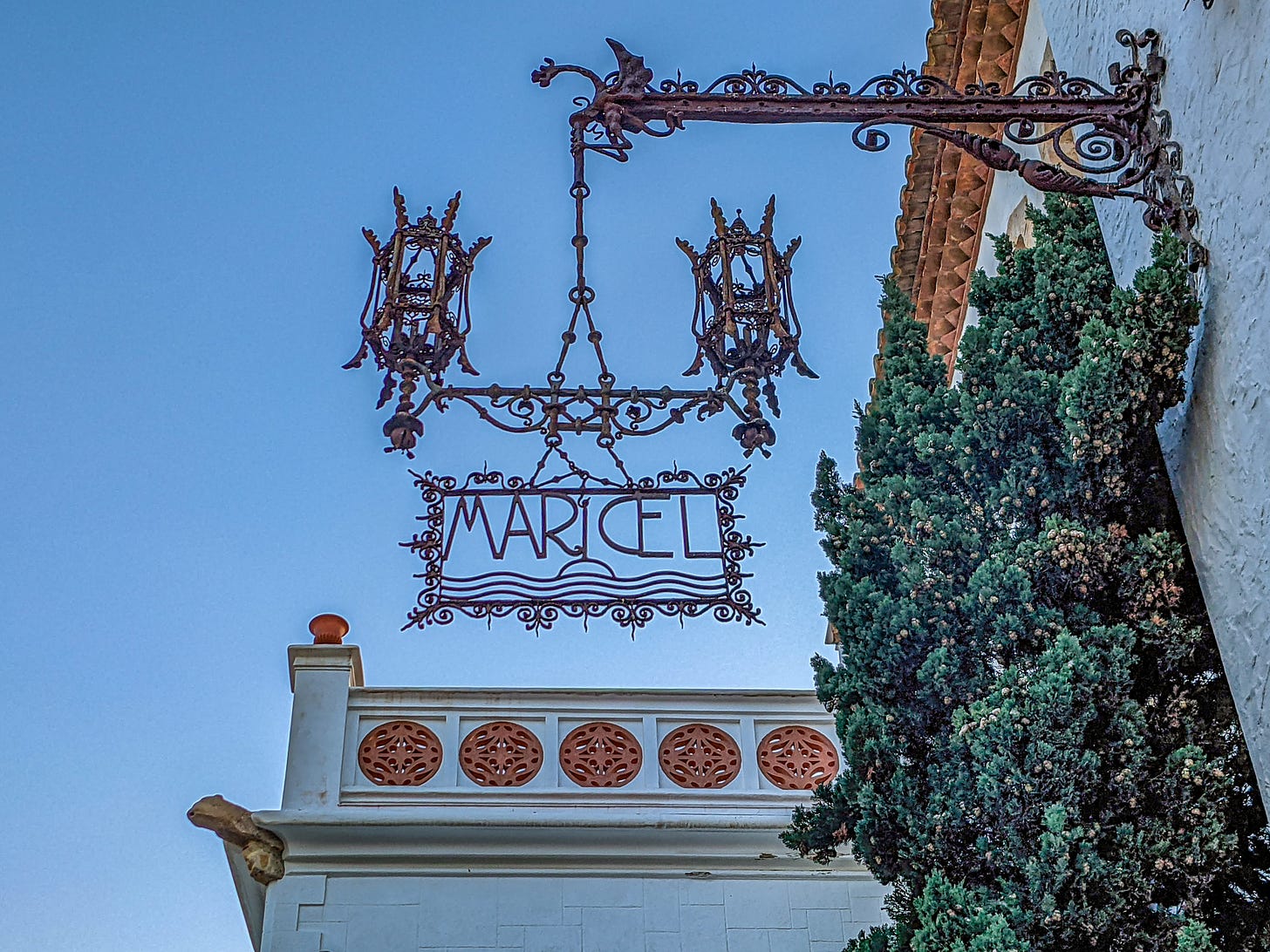 The elaborate wrought-iron sign for Museu de Maricel hanging from the side of a building and including two elaborate lamps. The elaborate wrought-iron sign for Museu de Maricel hanging from the side of a building and including two elaborate lamps.