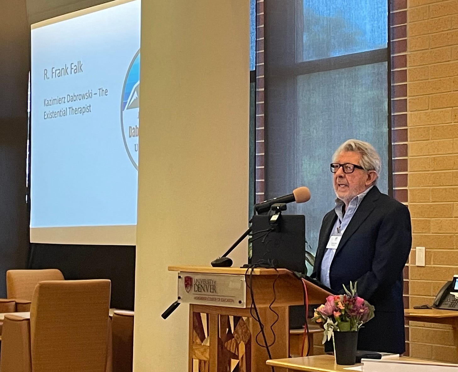 Frank Falk delivering his 2022 Dąbrowski Congress keynote from a University of Denver podium, wearing a dark blazer over a light blue shirt, speaking into a microphone with a small bouquet of flowers on the lectern.