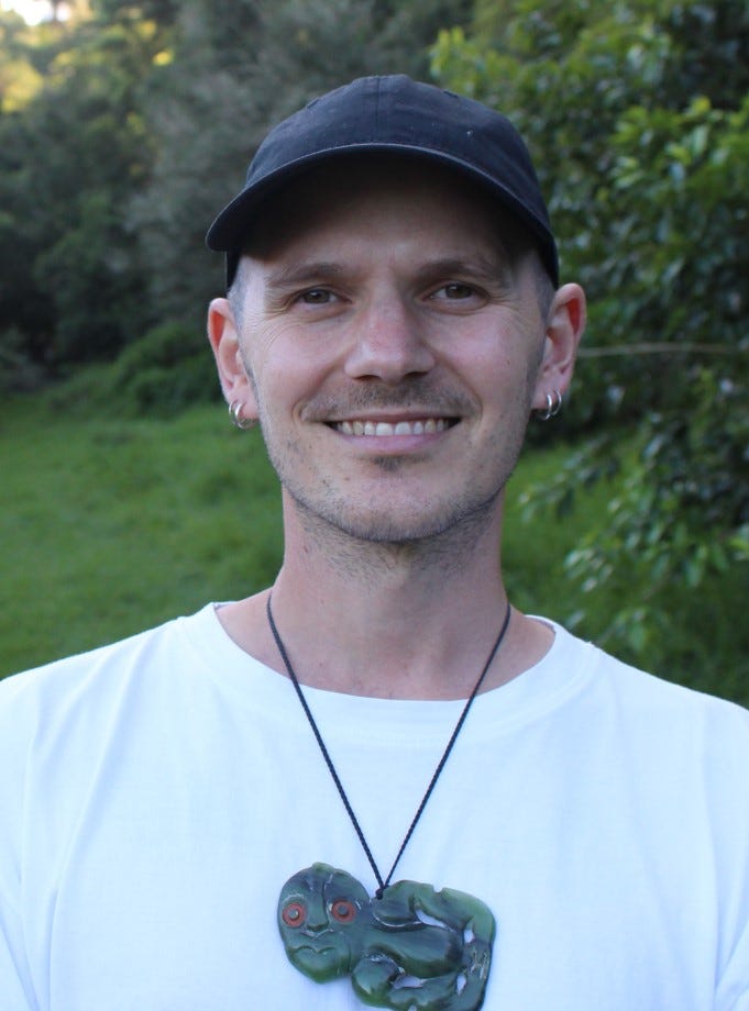 a person with a blue cap and white t-shirt, and several earings, a ponamu tiki around their neck
