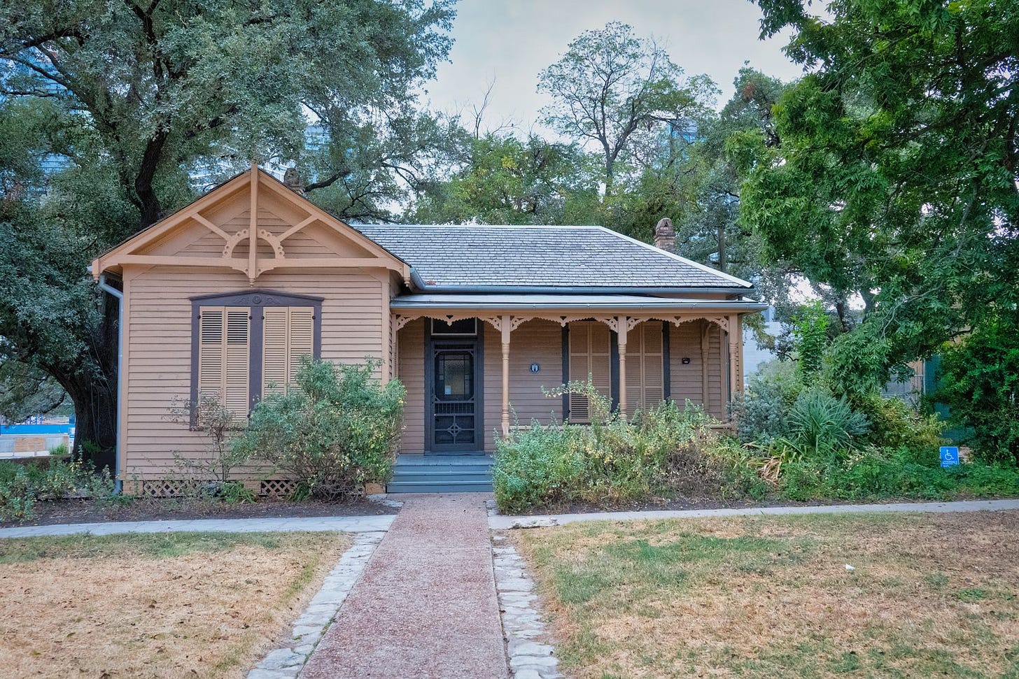 A single-storey wooden house with a covered front porch and an overgrown front yard, under mature trees A single-storey wooden house with a covered front porch and an overgrown front yard, under mature trees
