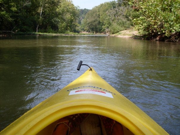 Front of kayak on river flanked by the trees.