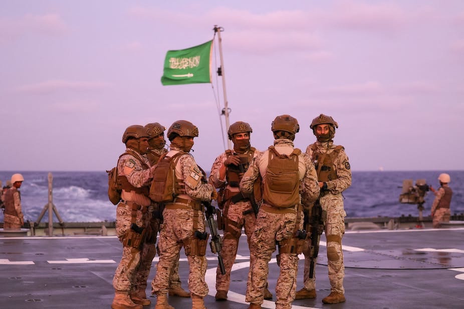 A group of men standing on board a military vessel with a flag hoisted on its mast. A group of men standing on board a military vessel with a flag hoisted on its mast.
