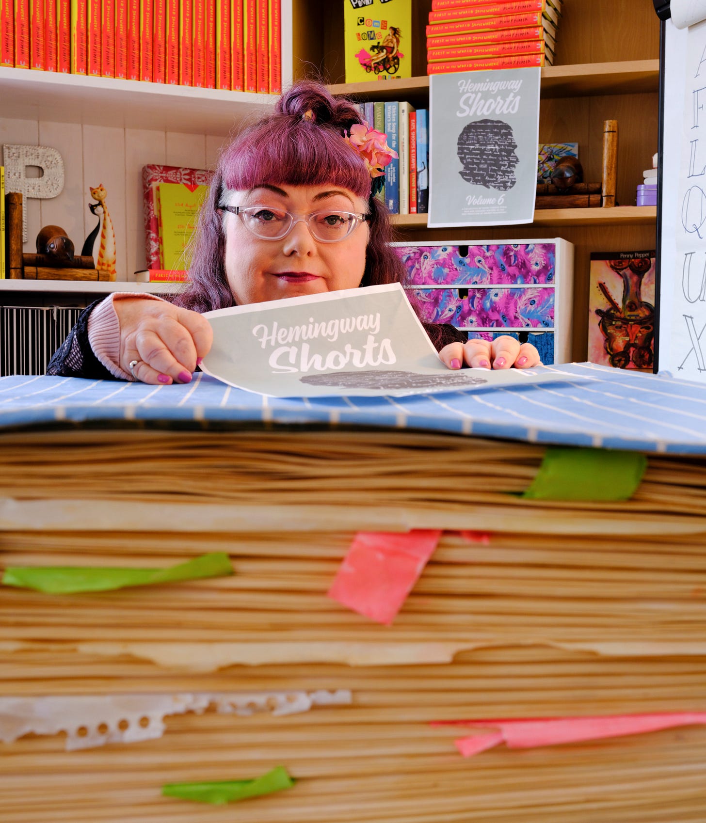 A photo of Penny in front of a giant diary prop used in her one-woman show, Lost in Spaces. The object in front of Penny represents her A5 diary from 1985 and shows a blue and white striped cover with cream-coloured pages crammed together, tatty Post-it notes and ripped notebook pages hanging between them. Penny is leaning on this prop, holding up a flier that says ‘Hemingway Shorts’. In the background are two bookcases showing Penny’s memoir, poetry collection and first edition of Desires. Penny has red hair and wears glasses. She is sitting in her wheelchair with a slight smile on her face.