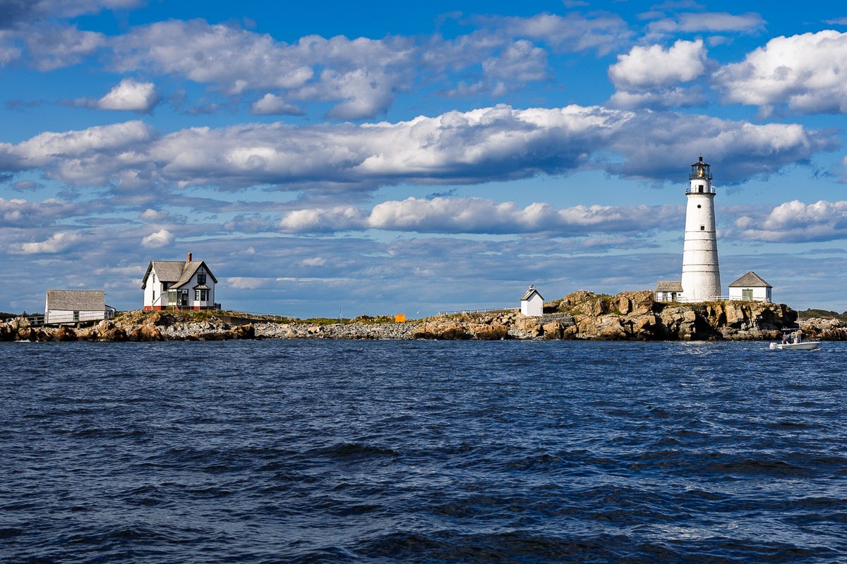 Boston Light lighthouse and keeper’s house on Little Brewster Island, seen from the water under blue sky and clouds, Boston Harbor