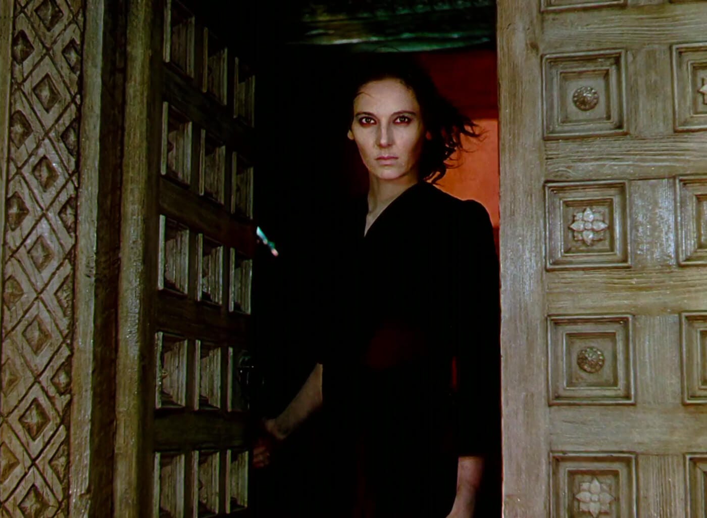 Sister Ruth stands in the convent doorway in her red dress, eyes fixed and hair wild, backlit by an infernal orange glow.