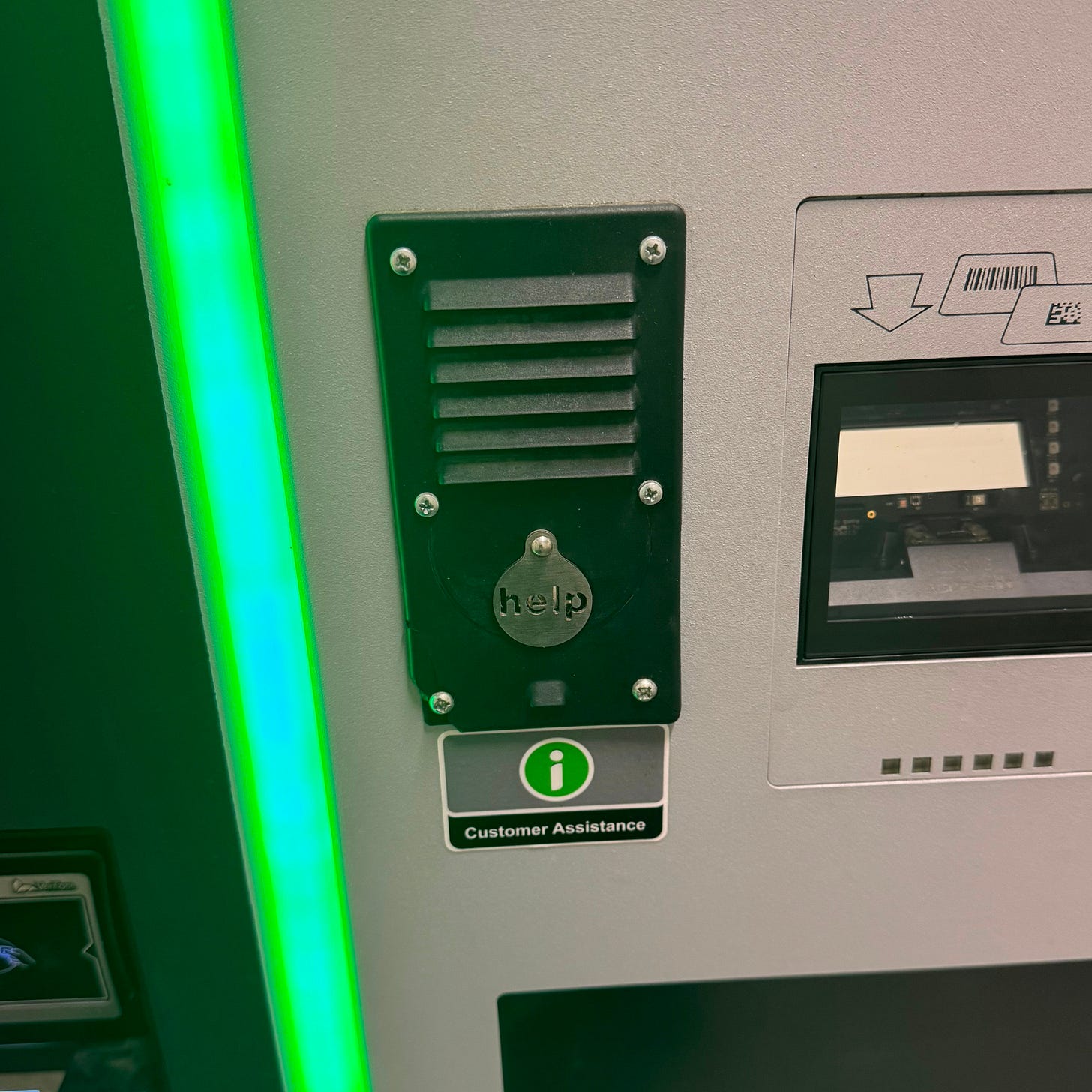 Close-up of a parking payment machine showing a black customer assistance intercom with a small silver button labeled ‘help,’ mounted next to a screen and beneath a green-lit edge.