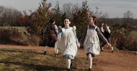 Two girls in 1890s clothing holding hands and running by the side of a school building