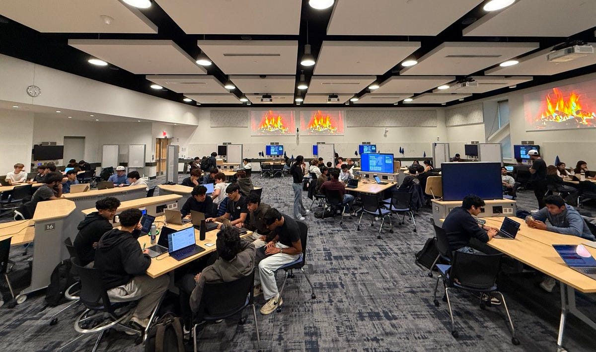A large modern classroom at University of Michigan with black ceiling panels and bright overhead lights features multiple rows of wooden desks occupied by over 100 young adults, mostly men in hoodies and casual attire, seated and working on laptops. Projector screens at the front and sides display orange flame graphics. A projector hangs from the ceiling, and large windows on the right side allow natural light. The floor is carpeted in gray, with no visible text or watermarks.