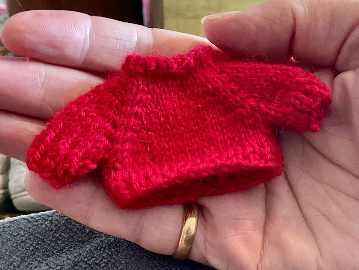 A red very tiny knitted pumper and a tiny knitted frog trying on two different types of shoes. He is green and wearing the red jumper and tartan shorts whilst sitting on a box to try on boots and shoes
