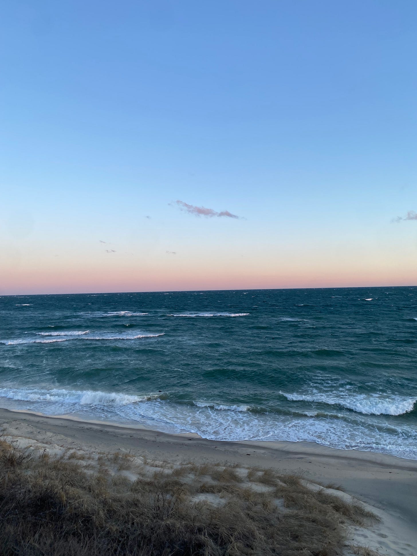 A beach at sunrise; white foamy waves under a blue sky with a pink-gold horizon.