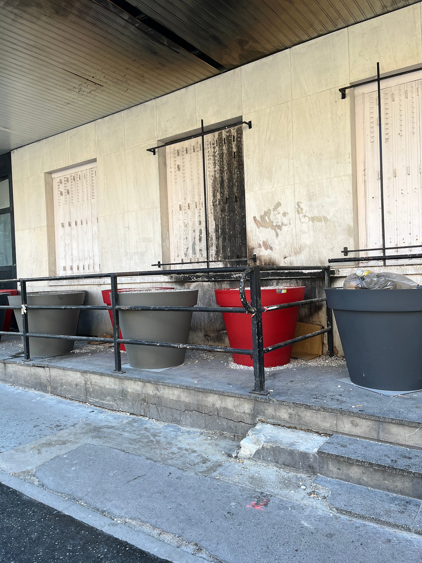 D'énorme pots de fleurs remplis de gravier poser sous l'auvent d'un immeuble. Ils sont sales, il y a du gravier au sol et des détritus dans un des pots.