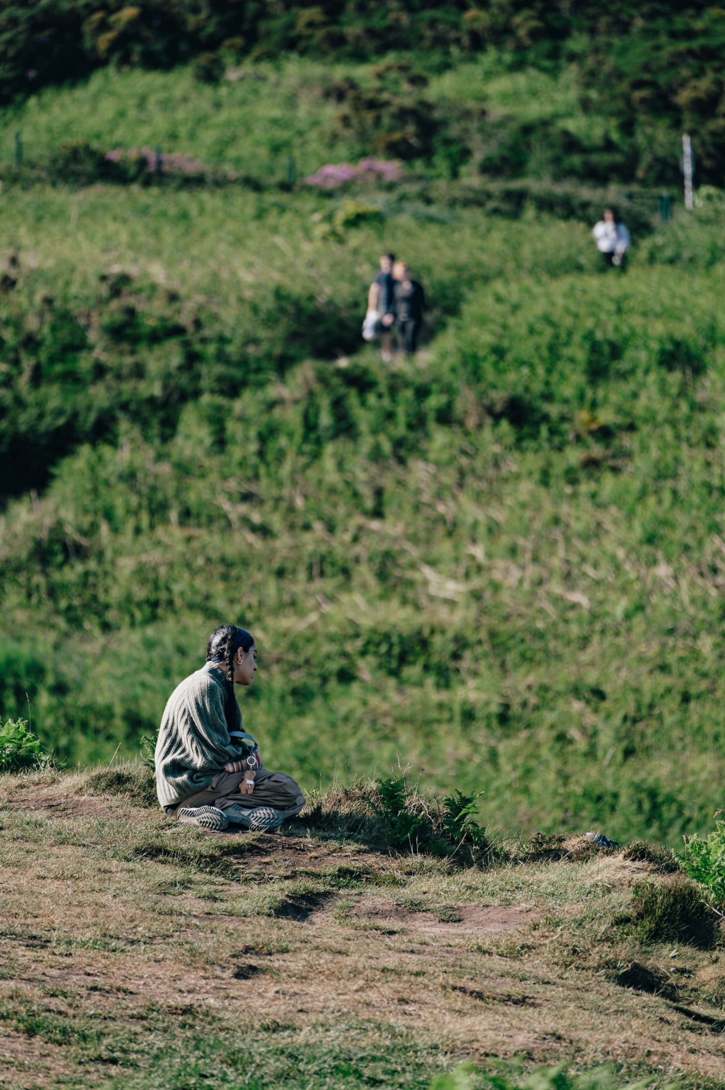 A solitary figure sits cross-legged on a coastal path, surrounded by green hills, gazing into the distance. A solitary figure sits cross-legged on a coastal path, surrounded by green hills, gazing into the distance.