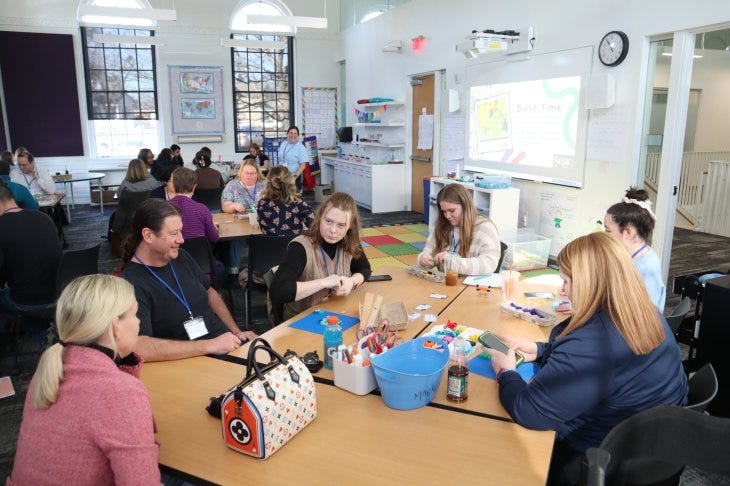 Participants sit in tables throughout a classroom, working with colorful objects.