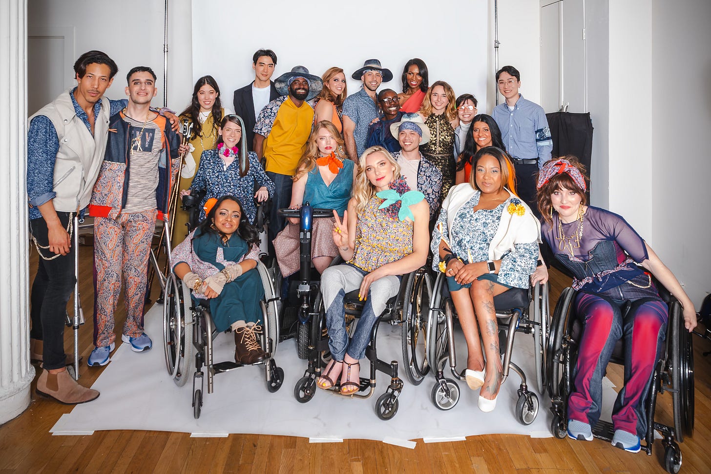 Full shot of the entire cast and crew of the Design for Disability project. About twenty diverse models and designers are gathered in a bright photo studio. In the front row, several women are seated in wheelchairs, smiling at the camera. Behind them, other models and designers are standing, some using crutches. Everyone is dressed in unique, colorful outfits from the fashion collection. Full shot of the entire cast and crew of the Design for Disability project. About twenty diverse models and designers are gathered in a bright photo studio. In the front row, several women are seated in wheelchairs, smiling at the camera. Behind them, other models and designers are standing, some using crutches. Everyone is dressed in unique, colorful outfits from the fashion collection.