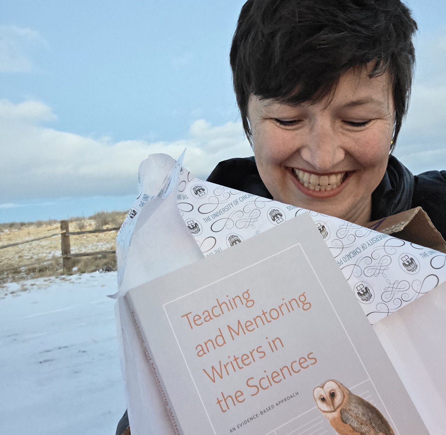 Smiling woman stands outdoors in a snowy environment. She is looking down at a book in her hands. The title of the book is legible: Teaching and Mentoring Writers in the Sciences.