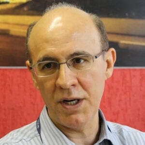Homem calvo de óculos falando, com camisa social de listras verticais azuis, tendo ao fundo a foto de uma rua colocada numa parede vermelha Homem calvo de óculos falando, com camisa social de listras verticais azuis, tendo ao fundo a foto de uma rua colocada numa parede vermelha