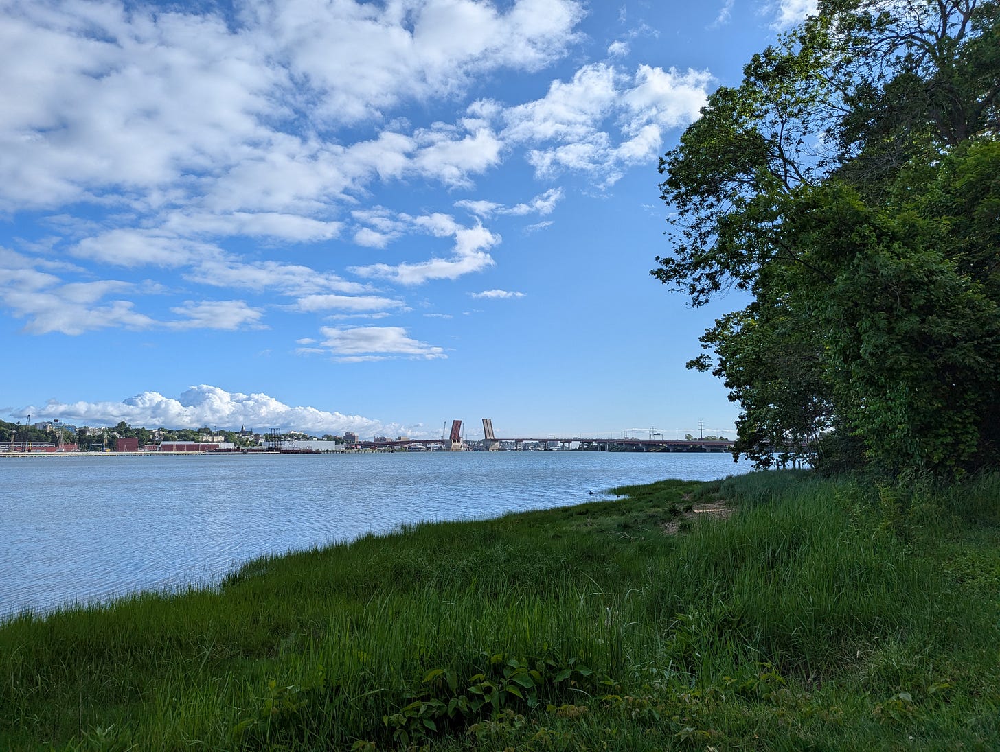 A grassy bank in the foreground, next to the high-tide waters of the Casco Bay in Portland, Maine, with the span of the drawbridge in the distance. The drawbridge is open. The sky is blue with fluffy white clouds. A grassy bank in the foreground, next to the high-tide waters of the Casco Bay in Portland, Maine, with the span of the drawbridge in the distance. The drawbridge is open. The sky is blue with fluffy white clouds.