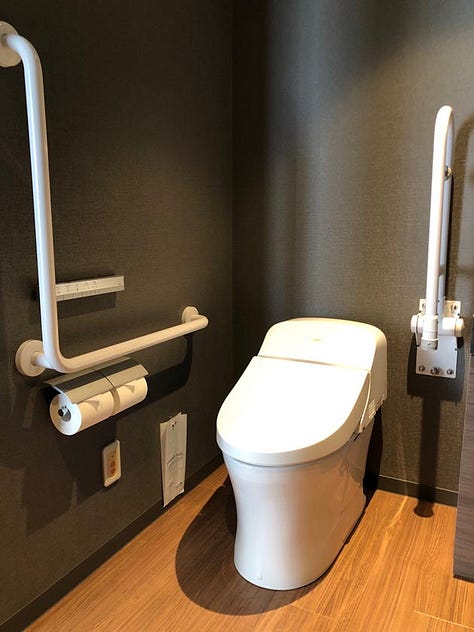 A modern and spacious hotel room with twin beds, a seating area with armchairs and a chaise lounge, a flat-screen TV, and large windows with sheer curtains. The accessible bathroom features grab bars, a handheld showerhead, and a shower chair.