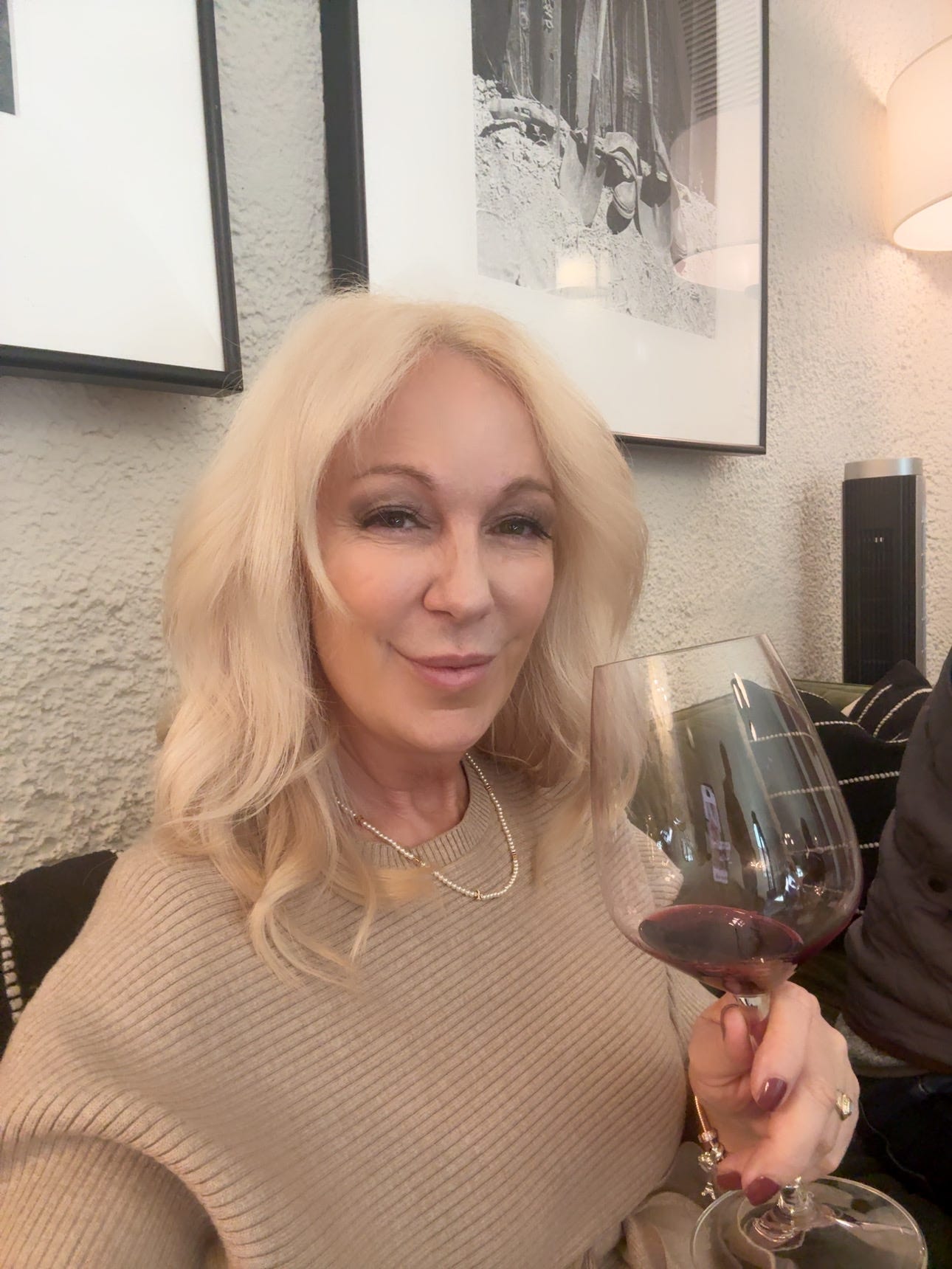 A woman with long blonde hair wearing a beige sweater is smiling while holding a glass of red wine.