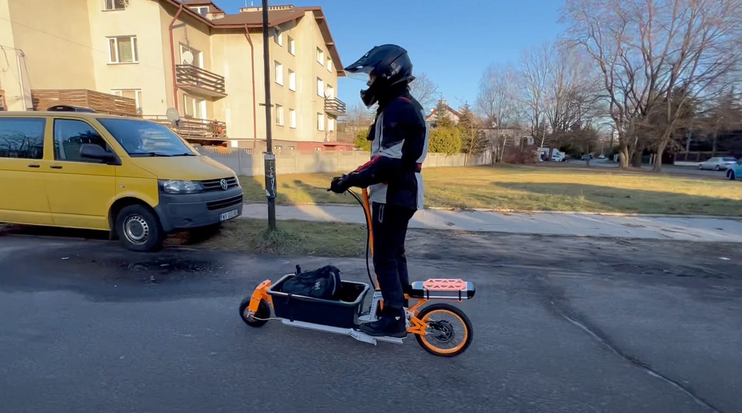 A man wearing motorcycle clothing and helmet rides on a small electric scooter. He stands towards the back of the scooter, and between him and the front wheel is a medium sized plastic box for carrying cargo.