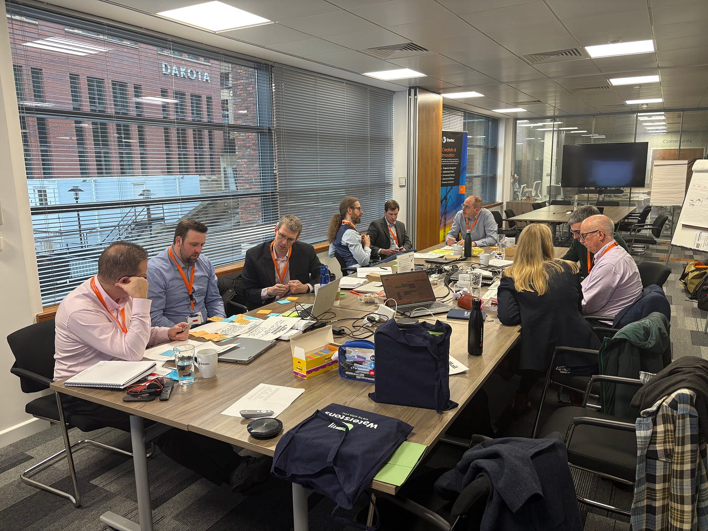 An office meeting room with slatted blinds and a large table. Around the table sit a group of men and women all wearing suits and orange lanyards. They are in groups, poring over a3 sheets covered in post its and sharpies. A workshop is in full swing.