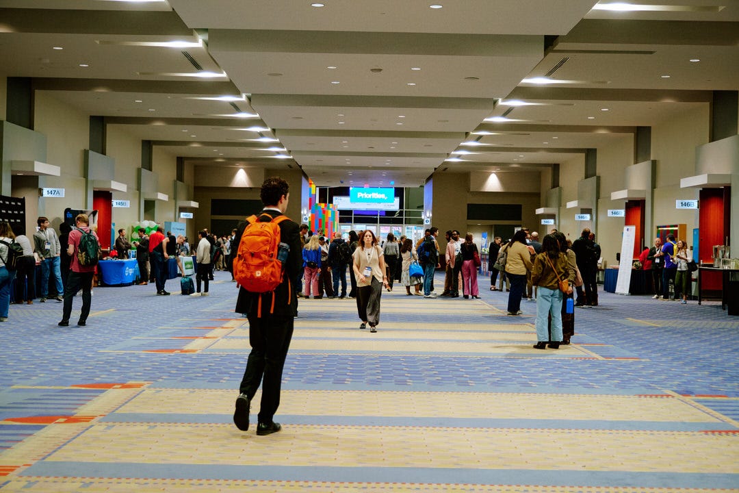 Wide shot down the long convention center hallway with the orange backpack guy in the foreground and hundreds of people moving between breakout rooms in the background