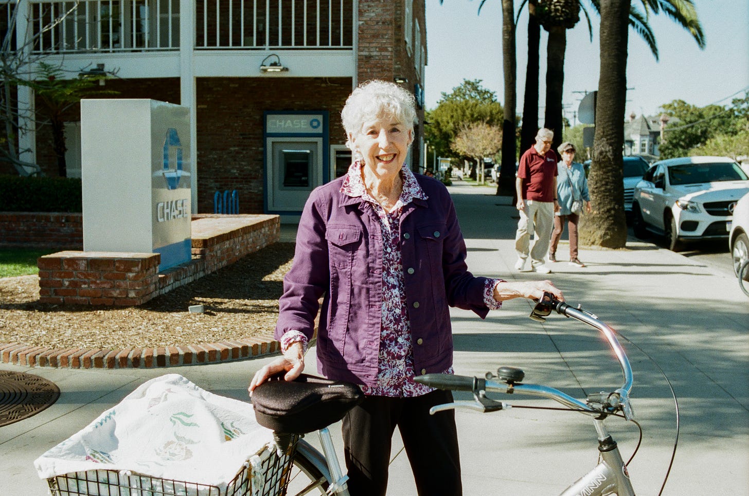 Older woman wearing purple with purple streaks in her hair, a three wheeled bike, and a dog in the basket. She's smiling.