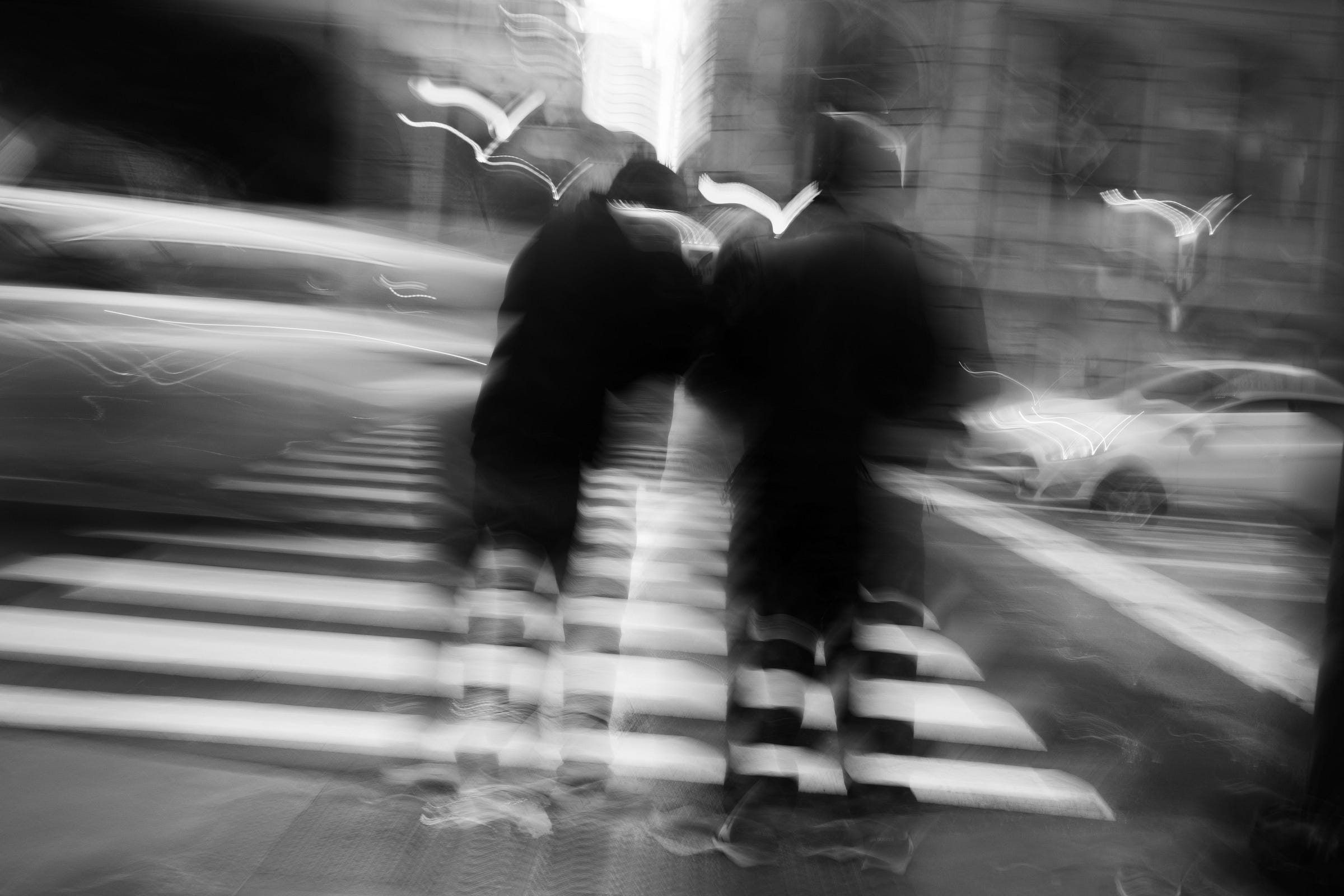 Two blurred figures cross a city street in motion, their forms blending with streaks of light from passing cars and distorted reflections on the wet pavement. Two blurred figures cross a city street in motion, their forms blending with streaks of light from passing cars and distorted reflections on the wet pavement.