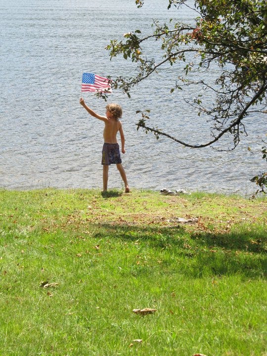 A small shirtless boy stands in shorts on grass, in front of a lake, waving a small American flag