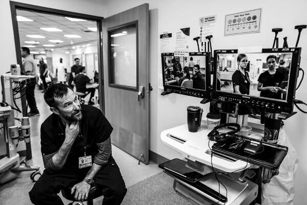 A black-and-white image of a bearded man in hospital scrubs in a hospital emergency department. He is watching doctors and patients elsewhere in the department on an incongruous array of television screens and audiovisual equipment. A black-and-white image of a bearded man in hospital scrubs in a hospital emergency department. He is watching doctors and patients elsewhere in the department on an incongruous array of television screens and audiovisual equipment.
