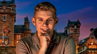 A young man in a brown shirt holds a hand to his chin as he smiles to camera, set against the backdrop of a castle by night A young man in a brown shirt holds a hand to his chin as he smiles to camera, set against the backdrop of a castle by night