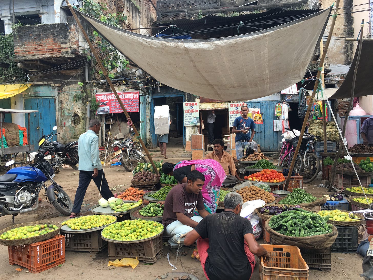 A vegetable stand is shown, with various bowls and crates full of gourds, tomatoes, carrots, potatoes, and the like. Two men and a women crouch down, while other men sit or walk in the background. A vegetable stand is shown, with various bowls and crates full of gourds, tomatoes, carrots, potatoes, and the like. Two men and a women crouch down, while other men sit or walk in the background.