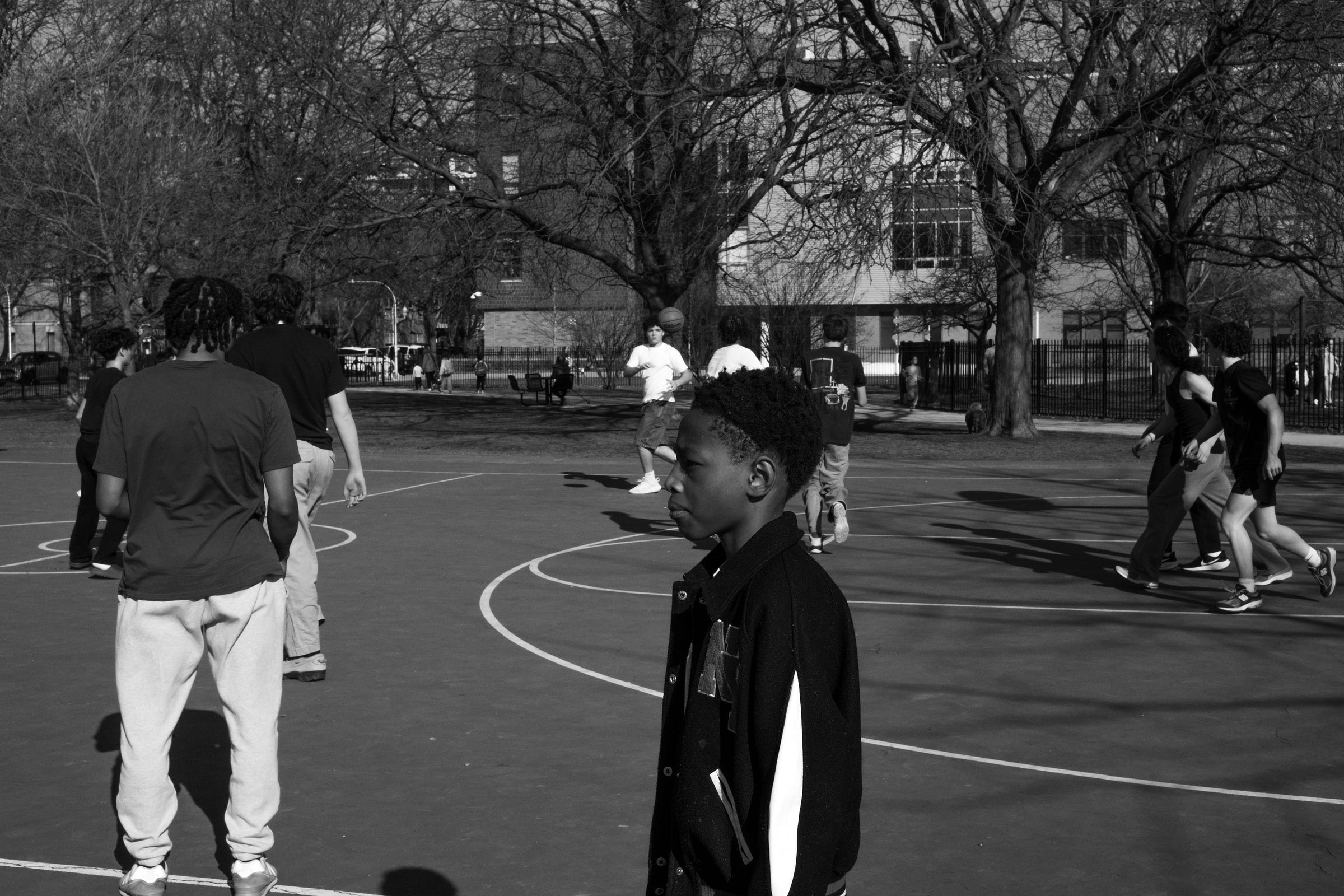 A black-and-white photograph of a young boy standing at the edge of a basketball court, gazing at the ongoing game with a contemplative expression. He wears a varsity-style jacket, contrasting with the more casually dressed players in the background. The court is filled with movement as older players run and pass the ball. Leafless trees and urban buildings frame the scene, adding depth and context to the environment. The boy's presence in the foreground suggests curiosity, anticipation, or a quiet moment of observation.