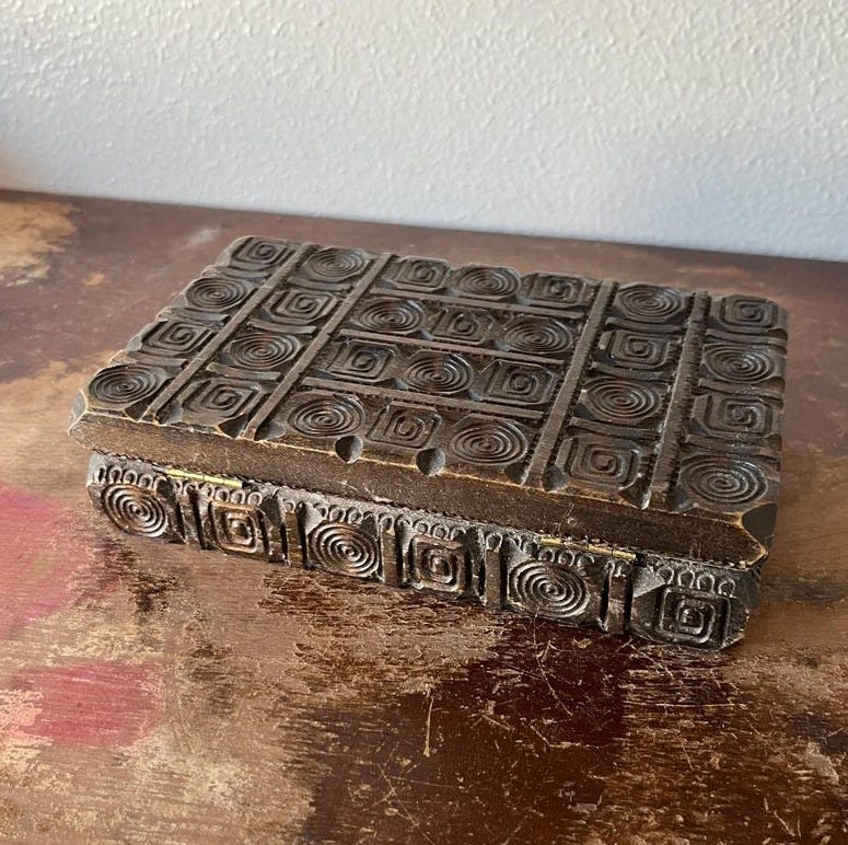 May include: A small, rectangular wooden box with a carved design. The box is dark brown and has a geometric pattern of circles and squares. The box is closed and sitting on a wooden surface.