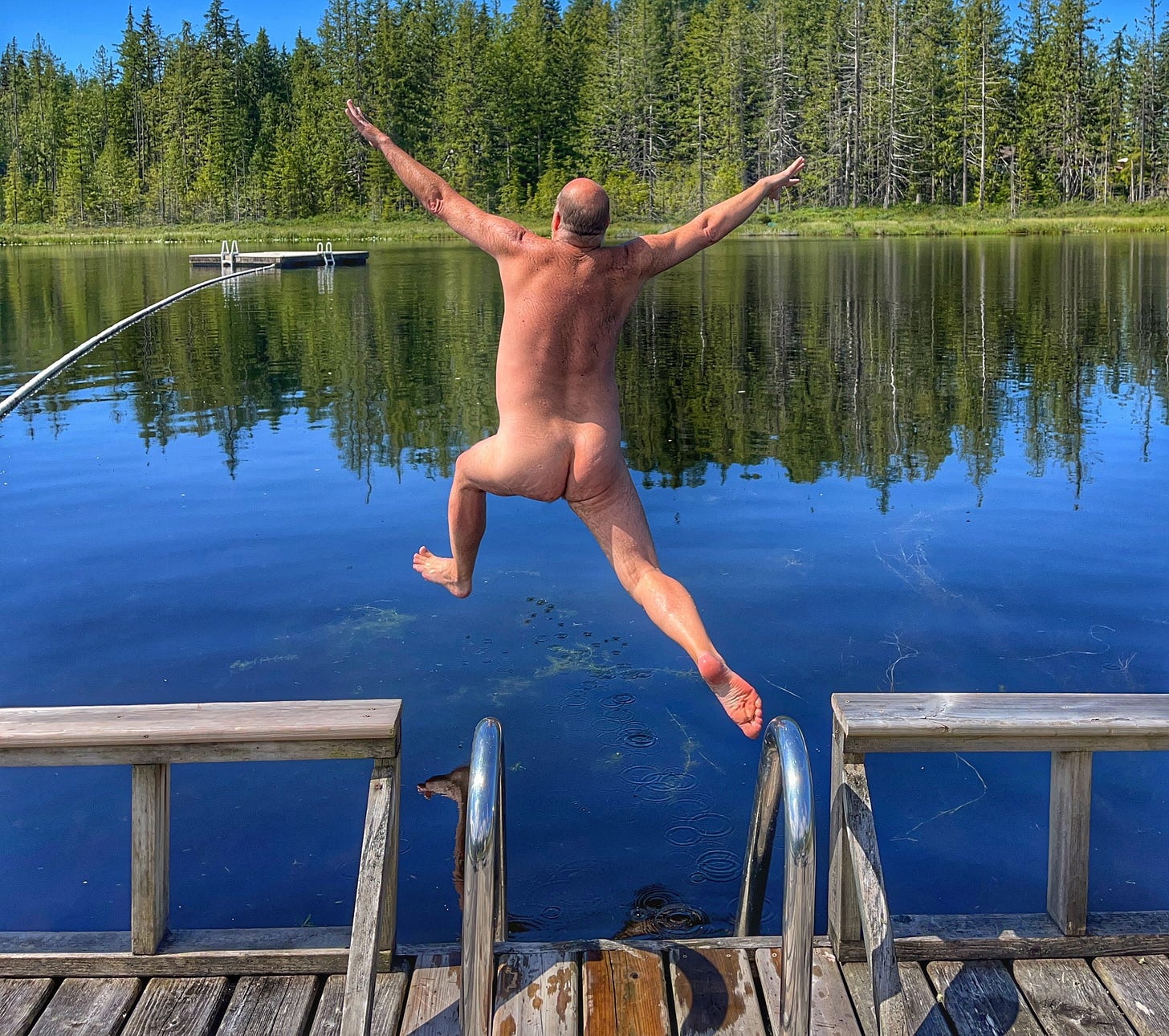 A nude man leaps energetically off a wooden dock into a clear, still lake surrounded by evergreen trees. His arms and legs are outstretched in midair as the calm water reflects the sky and forest. A nude man leaps energetically off a wooden dock into a clear, still lake surrounded by evergreen trees. His arms and legs are outstretched in midair as the calm water reflects the sky and forest.