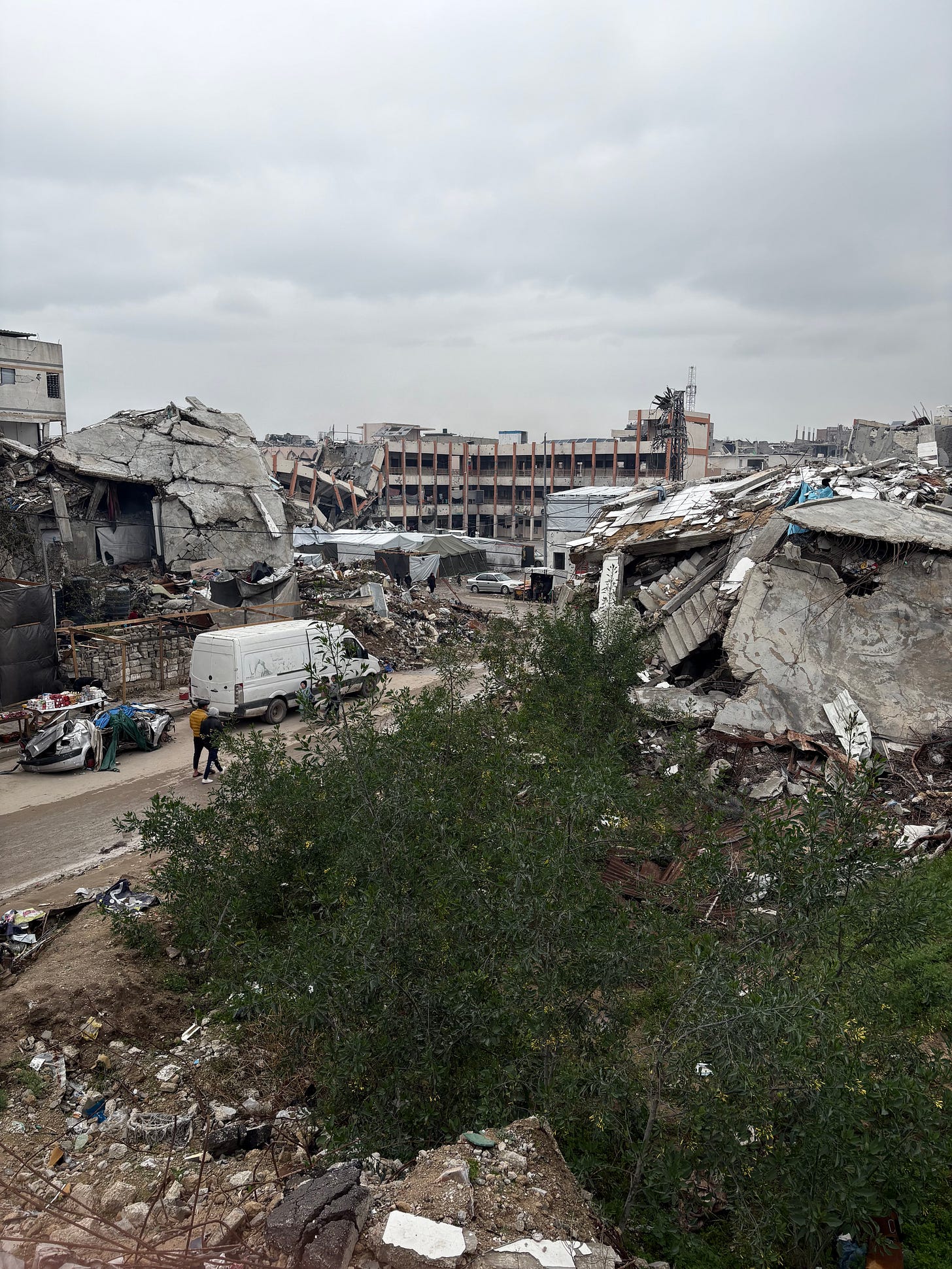 A wide view of a heavily damaged urban area with multiple large concrete buildings collapsed into piles of rubble under a gray, overcast sky. In the foreground, green shrubs and broken concrete frame the scene. A dirt road winds through the destruction, where a white van, a few cars, and several pedestrians are visible, highlighting ongoing activity despite the devastation. Makeshift shelters and scattered debris line the roadside, while a partially standing multi-story building rises in the background amid the ruins. A wide view of a heavily damaged urban area with multiple large concrete buildings collapsed into piles of rubble under a gray, overcast sky. In the foreground, green shrubs and broken concrete frame the scene. A dirt road winds through the destruction, where a white van, a few cars, and several pedestrians are visible, highlighting ongoing activity despite the devastation. Makeshift shelters and scattered debris line the roadside, while a partially standing multi-story building rises in the background amid the ruins.