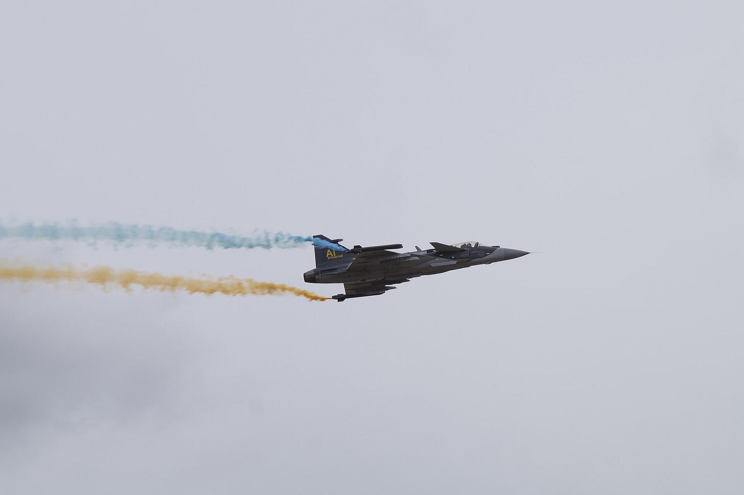 A Swedish Gripen E fighter banking against a grey sky, trailing blue and yellow smoke.