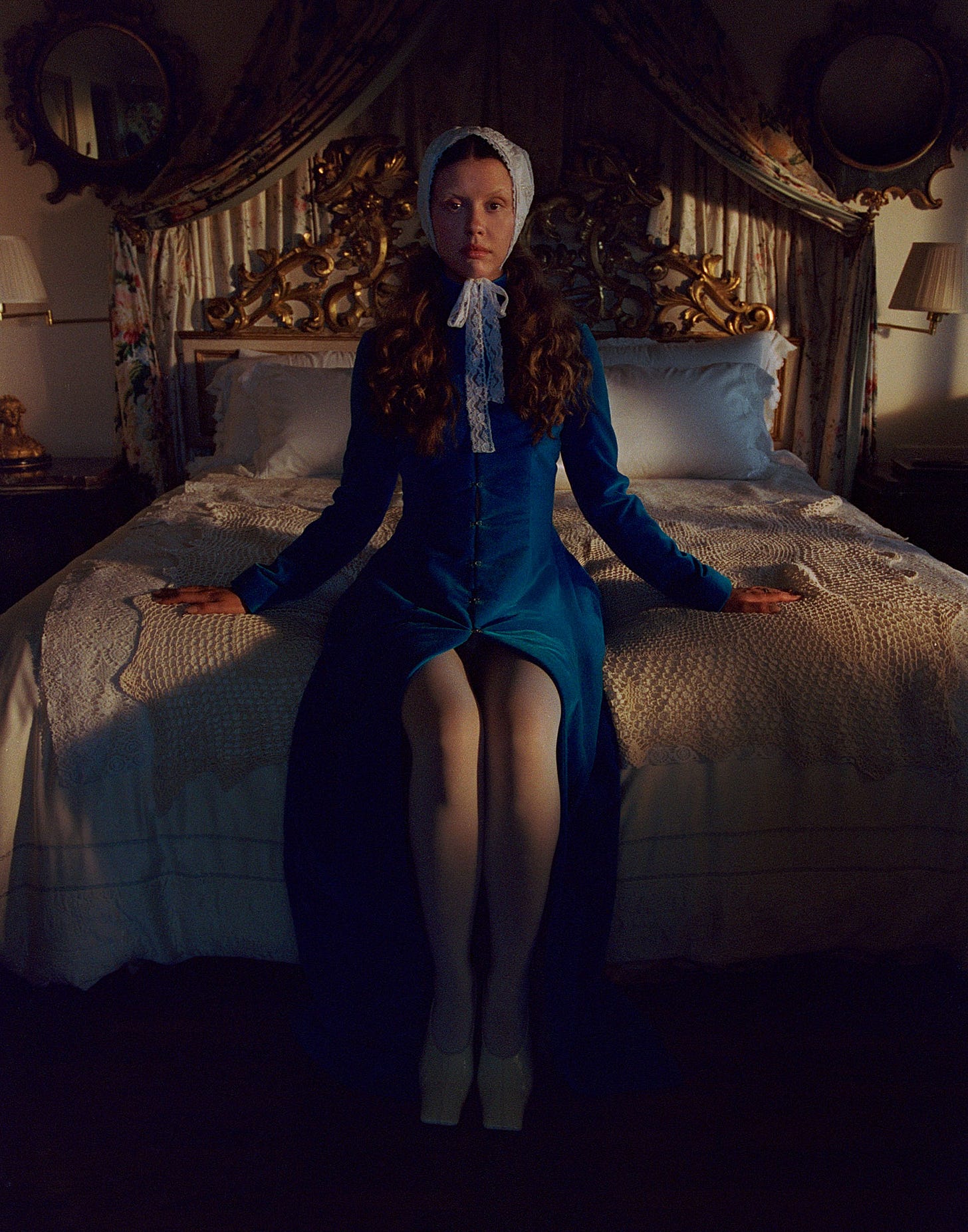 Mia Goth photo shoot for Who What Wear&#039;s October Cover Story. Mia Goth is sitting on a bed with ornate headboard. She is wearing a blue velvet long sleeve dress with white tights and a white lace headscarf.