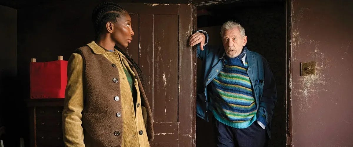 still from the christophers; an elderly white man stands in a run down doorway talking to a much younger Black woman