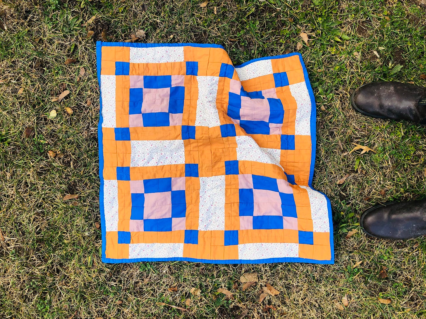 Photo of a small patchwork quilt with orange, pink, blue, and floral squares on grass with brown boots peeking into the frame