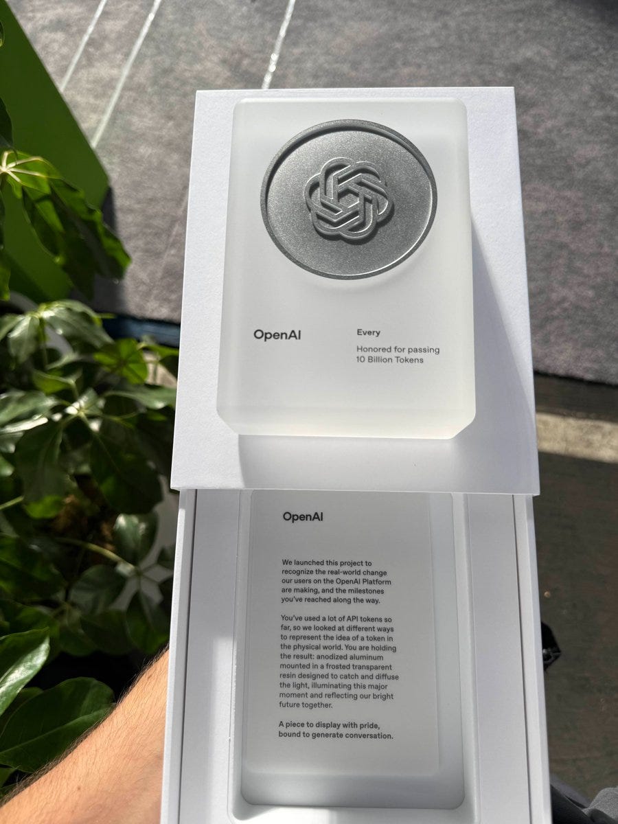 A white rectangular award plaque with a metallic hexagonal emblem featuring an interlocking knot design sits on a green surface outdoors with blurred greenery in the background. The plaque has OpenAI branding and text reading Honored for passing 10 Billion tokens. Below it, an open white box displays a printed letter on beige paper with OpenAI logo, text about opening new possibilities, changing the world, and future potential, signed with pride. A hand holds the box open.