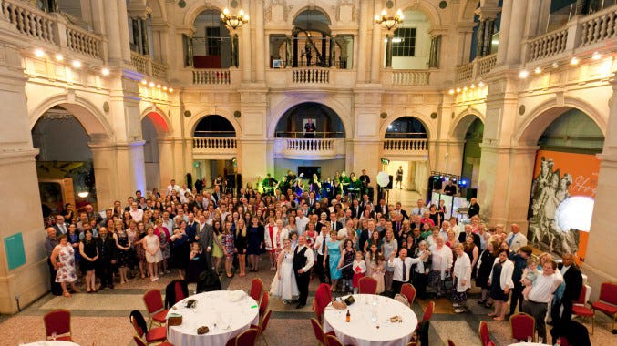 A large, diverse wedding party gathered in the grand, light-filled central hall of the Bristol Museum & Art Gallery, featuring high ceilings, ornate architecture, and museum exhibits in the background. The bride and groom are visible in the centre, surrounded by guests at round tables set for a reception, illustrating the "unique marriage venue" trend discussed on www.thealmightygob.com. A large, diverse wedding party gathered in the grand, light-filled central hall of the Bristol Museum & Art Gallery, featuring high ceilings, ornate architecture, and museum exhibits in the background. The bride and groom are visible in the centre, surrounded by guests at round tables set for a reception, illustrating the "unique marriage venue" trend discussed on www.thealmightygob.com.