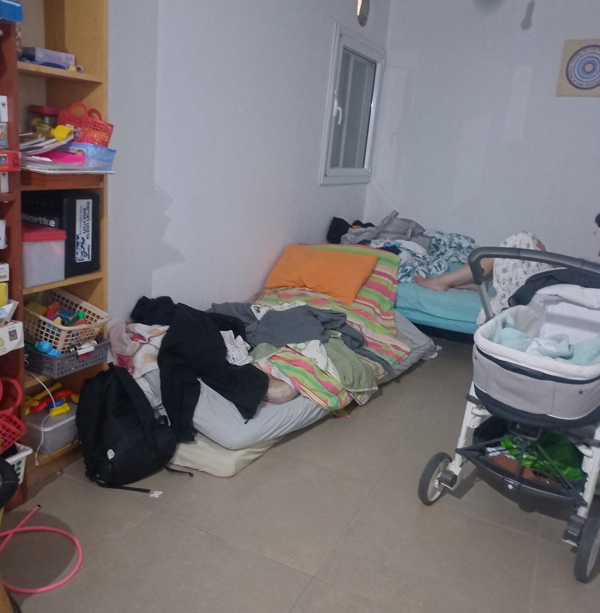 Interior of a safe room with shelves of toys, a pile of mattresses, a baby carriage, and partial view of someone sitting on the bed