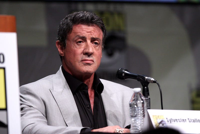 A man in a black shirt and light gray suit jacket, sitting behind a microphone and bottle of water A man in a black shirt and light gray suit jacket, sitting behind a microphone and bottle of water