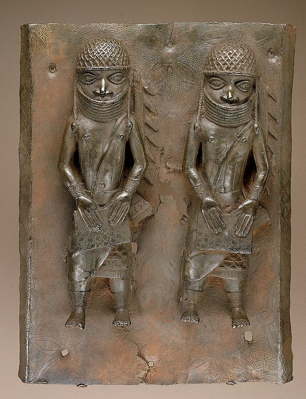 Bronze plaque with two raised figures in matching attire. They wear helmet-like head coverings, neck ornamentation that covers their mouths,  and straps that cross their chest diagonally, perhaps connected to a bag or satchel. 