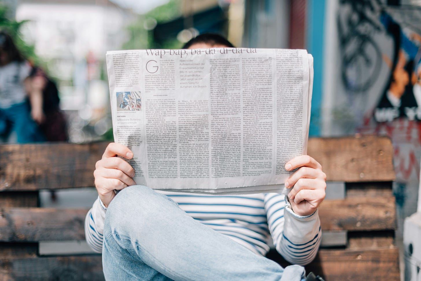 A man sits on a wooden bench, holding up a newspaper in front of his face.