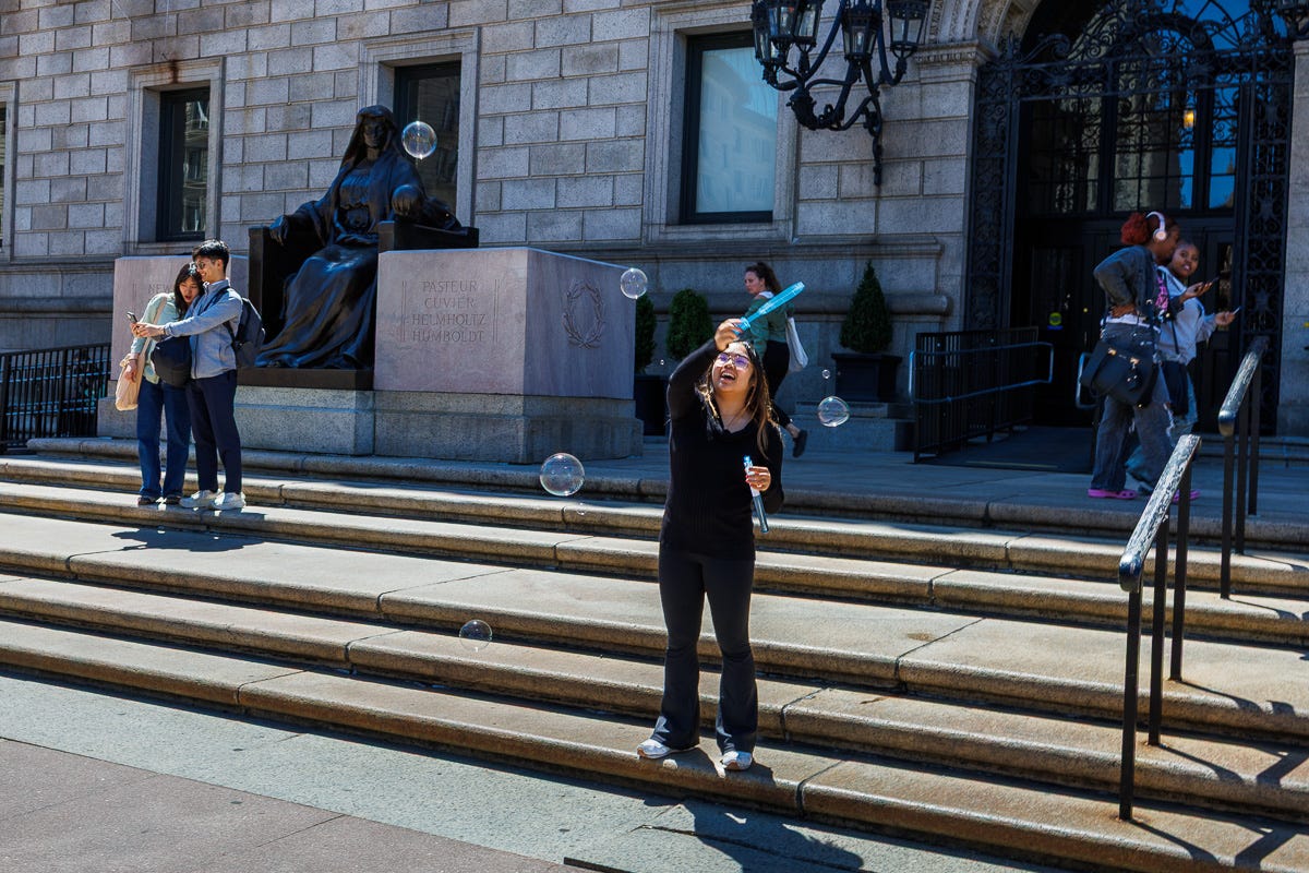A woman joyfully blows bubbles on the steps of the Boston Public Library as others take selfies nearby in the sunshine. A woman joyfully blows bubbles on the steps of the Boston Public Library as others take selfies nearby in the sunshine.