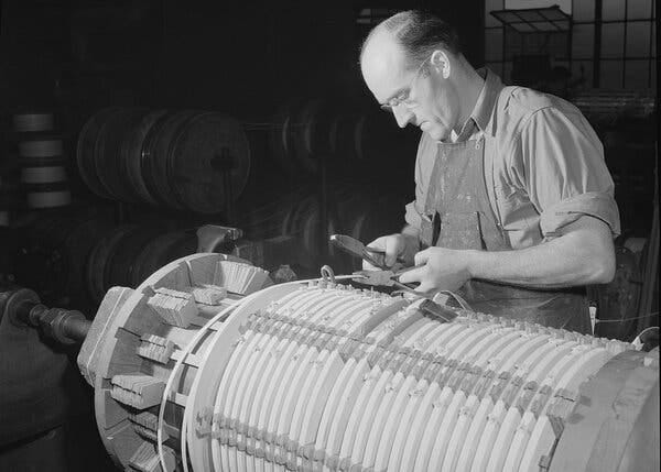 A black-and-white photo of a person using pliers to wind coils around a similar-looking large cylindrical piece of equipment. A black-and-white photo of a person using pliers to wind coils around a similar-looking large cylindrical piece of equipment.