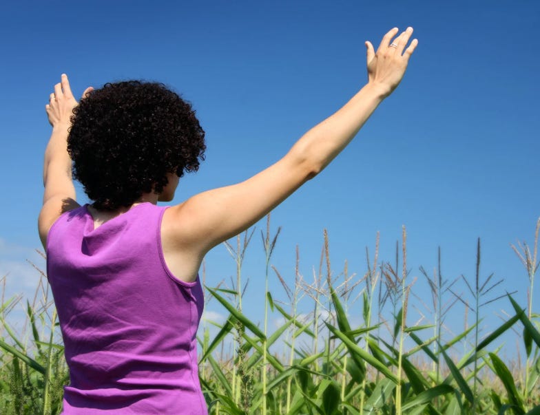 Woman with hands raised to the sky in praise while standing in a corn field.