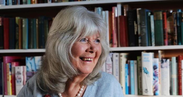An image of Jilly Cooper, an older white woman with grey hair. She is standing in front of a full bookcase, smiling and wearing a string of pearls and a pale blue cardigan.